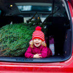 Adorable little toddler girl with Christmas tree inside of family car. Happy healthy baby child in winter fashion clothes choosing and buying big Xmas tree for traditional celebration.