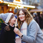 Mother and daughter eating white chocolate covered fruits and strawberry on skewer on traditional German Christmas market. Happy girl and woman on traditional family market in Germany during snowy day
