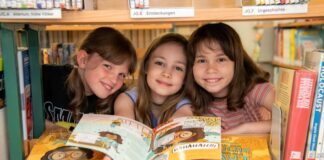 Drei Mädchen lächeln in die Kamera und zeigen ein geöffnetes Heft her.