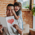 Smiling child hiding father's eyes and handing gift postcard